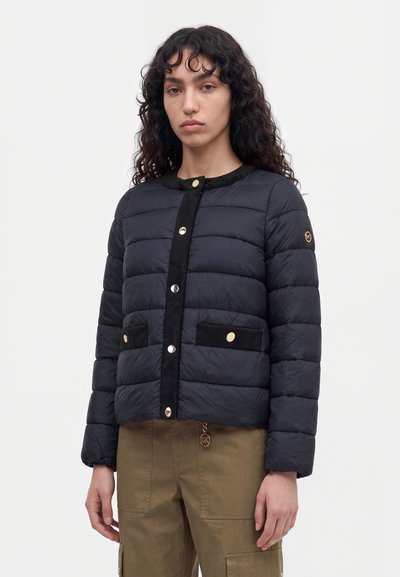 Une femme porte une veste matelassée noire à boutons dorés et un pantalon cargo marron, sur un fond blanc uni.