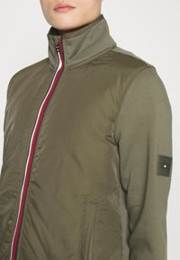 Kurtka bombowiec w oliwkowej zieleni z wysokim kołnierzem, dwukolorowym zamkiem błyskawicznym w czerwono-białe pasy oraz naszywką z logo na rękawie. Gładka faktura.