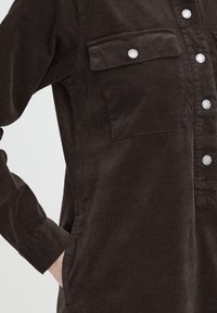 Krupni plan tamnosmeđe manšestarske košulje sa srebrnim kopčama na pritisak i džepom na prsima, ruka smještena u bočni džep.