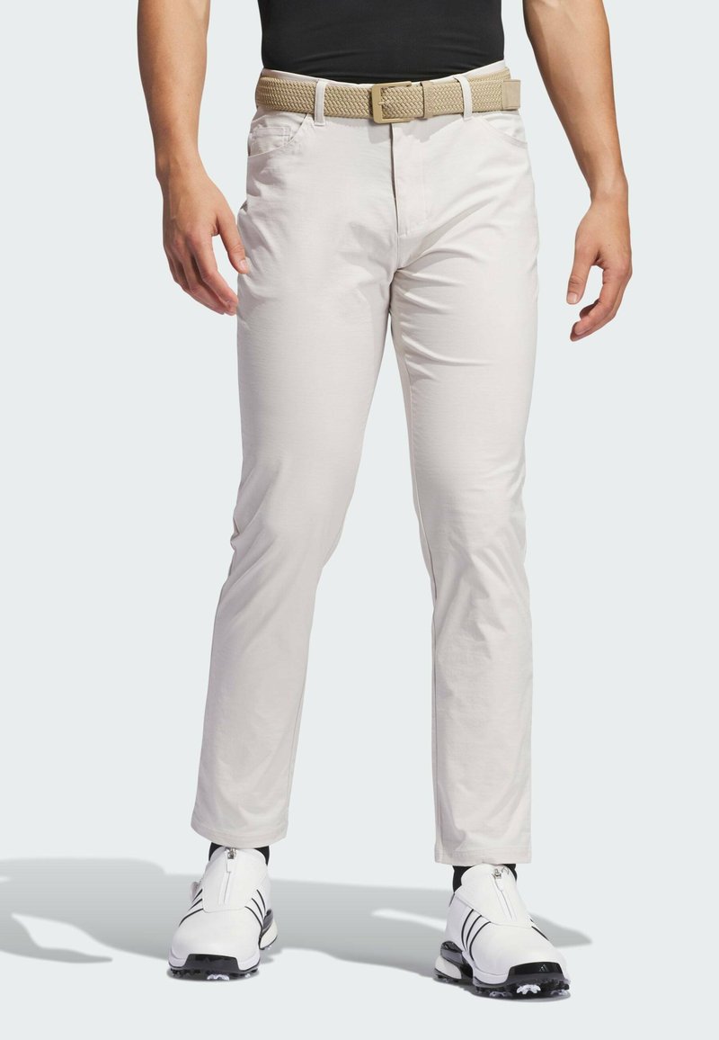 Chinos de color beige claro con una textura suave, corte recto y bolsillos laterales, acompañados de un cinturón trenzado marrón claro y zapatos de golf blancos.