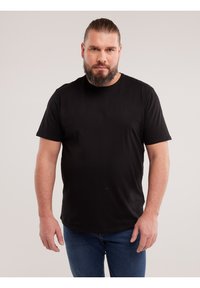 Homme adulte avec une barbe et de longs cheveux attachés, portant un t-shirt noir et un jean bleu, debout devant un fond clair uni.