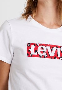 T-shirt bianco in cotone con un logo rettangolare a stampa leopardo rossa e la parola "Levi's" in lettere bianche in grassetto. Design classico con scollatura a girocollo.