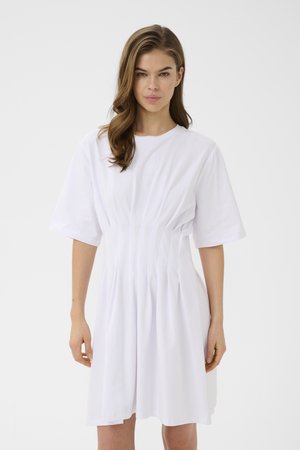 Kvinde med bølget brunt hår iført en hvid, plisseret, kortærmet midikjole, stående mod en ensfarvet hvid baggrund.
