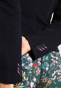 Dark navy blazer features textured fabric, pink stitching details, and three black buttons at the cuff, paired with floral-patterned trousers.