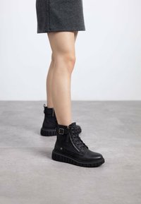 Botas negras hasta el tobillo de cuero con suela texturada, cordones al frente, cremallera lateral y acento de hebilla metálica. Acabado suave, diseño resistente.