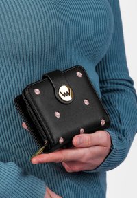 Black leather wallet with pink polka dots, a gold logo clasp, and a zip closure detail, held in a hand against a textured blue background.