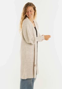 Glunderende vrouw in een beige lange cardigan over een zwart top en blauwe jeans, die haar mouw aanpast tegen een neutrale witte achtergrond.
