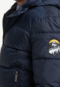 Marinblå pufferjacka med dragkedjestängning, strukturerad yta och en logotyp-patch på ärmen. Har en huva och ribbade detaljer.