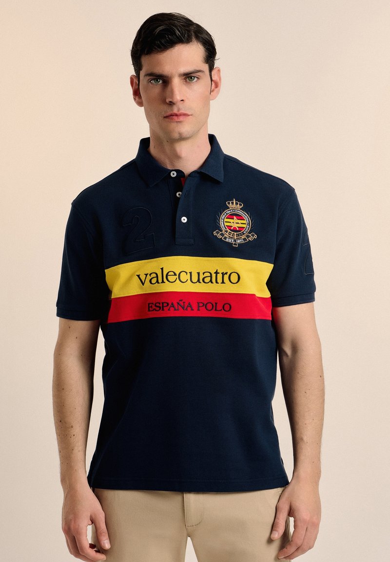 Polo navy con rayas horizontales amarillas y rojas, presenta un logo, el número 2 bordado y el texto "valecuatro" y "ESPAÑA POLO."