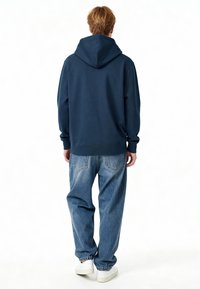 Felpa con cappuccio blu navy, dal fit rilassato, in cotone, con polsini a coste, indossata con jeans baggy azzurri e sneaker bianche.