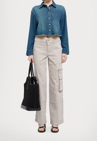 Camicia di jeans tagliata in blu scuro con orlo frangiato, abbinata a pantaloni cargo a gamba larga beige chiaro con grandi tasche. Inclusa borsa a rete nera.