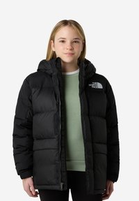 Melna pūkā jacket ar kapuci; ar quiltētu dizainu, ar rāvējslēdzēju priekšā un logo uz krūtīm; uzvilkta virs gaiši zaļa džempera.