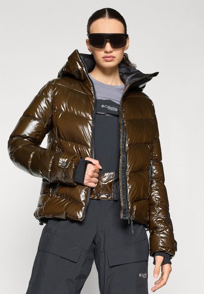 Barna puff-jackets fényes felülettel, cipzárral és kapucnival. Sötét felsőre van húzva, és sötét cargo nadrággal van párosítva. Rugalmas mandzsettákkal rendelkezik.