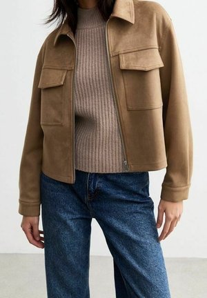 Femme portant une veste en daim beige avec fermeture éclair et poches sur la poitrine, un pull côtelé beige et un jean bleu, debout avec une main détendue.