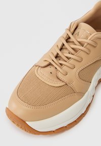 Barna sneaker, hálós és bőr felsőrésszel, bézs fűzőkkel, fehér középtalppal és gumi külsőtalppal, részletes varrással és szellőzőnyílásokkal.