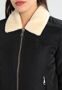 Giacca nera con zip, dotata di un colletto in pelliccia sintetica color crema, tasca laterale con zip e una texture di tessuto liscia con un leggero lucido.