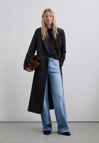 Lange geruite jas in donkere tinten, gecombineerd met een donker gemêleerd sweater en lichtblauwe wijde jeans. Houdt een bruine clutch vast.