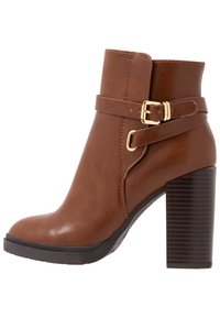 Hnědá kožená kotníková bota se zlatou přezkou, předním páskem a zaoblenou špičkou. Má robustní, vroubkovaný vysoký podpatek.