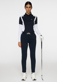 Navyblå pikétröja med vita detaljer, långärmad, tillsammans med skräddarsydda marinblå byxor. Håller en golfklubba, bär en vit handske och sneakers.