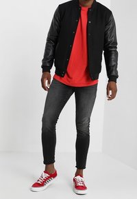 Chaqueta bomber negra con mangas de cuero sobre una camiseta roja, combinada con jeans negros ajustados y zapatillas rojas con un patrón de tablero de ajedrez negro.
