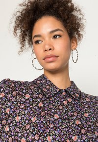 Blusa floral com fundo preto, apresentando flores multicolores, um colarinho e ombros bufantes. A modelo usa brincos de argola prateados.