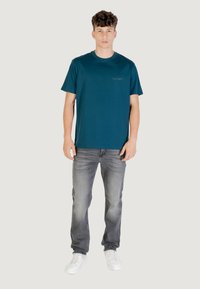 T-shirt in cotone verde acqua con collo a giro, maniche corte e un piccolo logo sul petto, abbinato a jeans in denim grigio sbiadito e sneaker bianche.