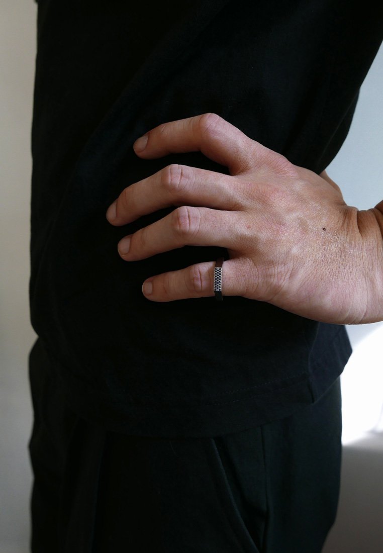 Melnā gredzena ar fakturētu, rakstainu virsmu, kas valkāts uz rokas, kas balstās uz tumšas apģērba. Kopējais izskats ir minimālisma un mūsdienīgs.