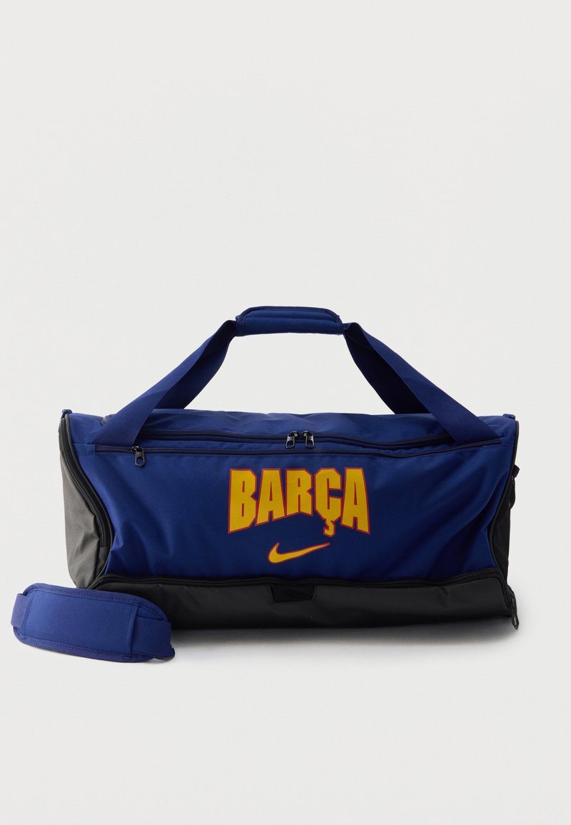 Μπλε και μαύρη τσάντα Nike duffel με κίτρινο κείμενο "BARÇA" στο μπροστινό μέρος και αφαιρούμενο επενδυμένο ιμάντα ώμου τοποθετημένο δίπλα της.
