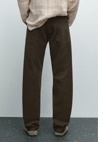Hnědé kalhoty rovného střihu vyrobené z bavlněného denimu. Mají dvě zadní kapsy s prošívanými detaily. Nosič má na sobě světle kostkovanou košili.