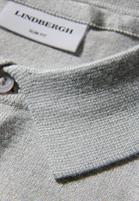 Zbliżenie na jasnoszarą koszulkę polo z fakturowaną tkaniną, guzikiem i przeszywanym kołnierzem. Metka z napisem "Lindbergh Slim Fit".