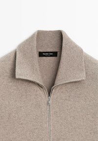Hellbeige Zip-Up-Pullover mit geripptem Kragen und Ärmeln, aus weichem Strickmaterial gefertigt, mit einem silbernen Reißverschlussdetail auf der Vorderseite.