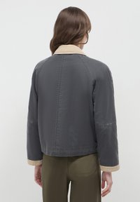 Giacca grigia corta con colletto e polsini beige, caratterizzata da una texture liscia e maniche raglan. Dettagli delle cuciture visibili sul retro.