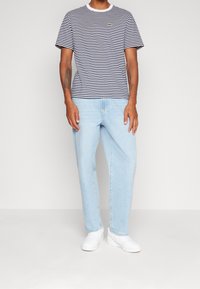 Man wearing navy and white striped short-sleeve shirt, light blue jeans, and white sneakers, standing against a plain light background.