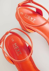 Orange högklackade sandaler med korsade remmar, texturerad konstläderöverdel, mjuk sula och guldvarumärke som är tryckt invändigt.