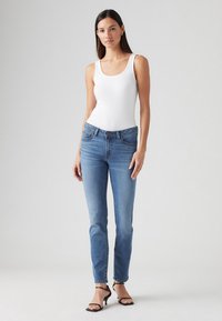 Kék skinny farmernadrág, enyhe elhalványulással, fehér pántos top-pal párosítva. A modellen fekete pántos magassarkú cipő van. Egyszerű dizájn, tiszta vonalak, minták nélkül.