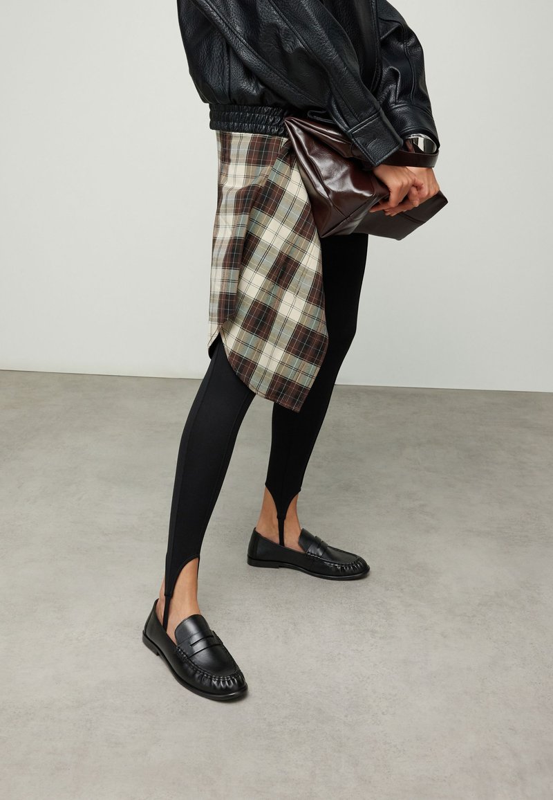 Zwarte leren jas, geruite wikkelrok, zwarte legging met uitsparingen en zwarte loafers. Accessoire: donkerbruine clutch.