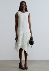 Rochie albă asimetrică cu un top structurat și tiv cu franjuri, asortată cu pantofi negri cu toc și o poșetă mică neagră. Textură netedă a materialului.