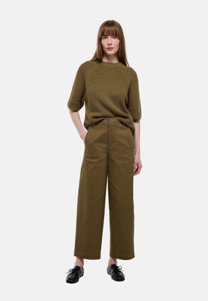 Femme portant un pull côtelé vert olive à manches trois-quarts, pantalon kaki à jambe large, et chaussures noires à lacets, debout avec une main dans la poche.