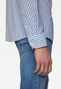 Blau-weiß gestreiftes Langarmshirt mit Knopfmanschetten, aus Stoff gefertigt, neben ausgewaschenen blauen Jeans präsentiert, mit seitlicher Ansicht.