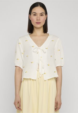 Vrouw met steil donker haar, gekleed in een wit kortemouwenblouse met kleine gele bloemembroderie en een lichtgele rok.