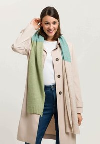 Écharpe en tricot avec un design dégradé en vert clair et turquoise, au toucher doux, drapée de manière décontractée sur un manteau crème.