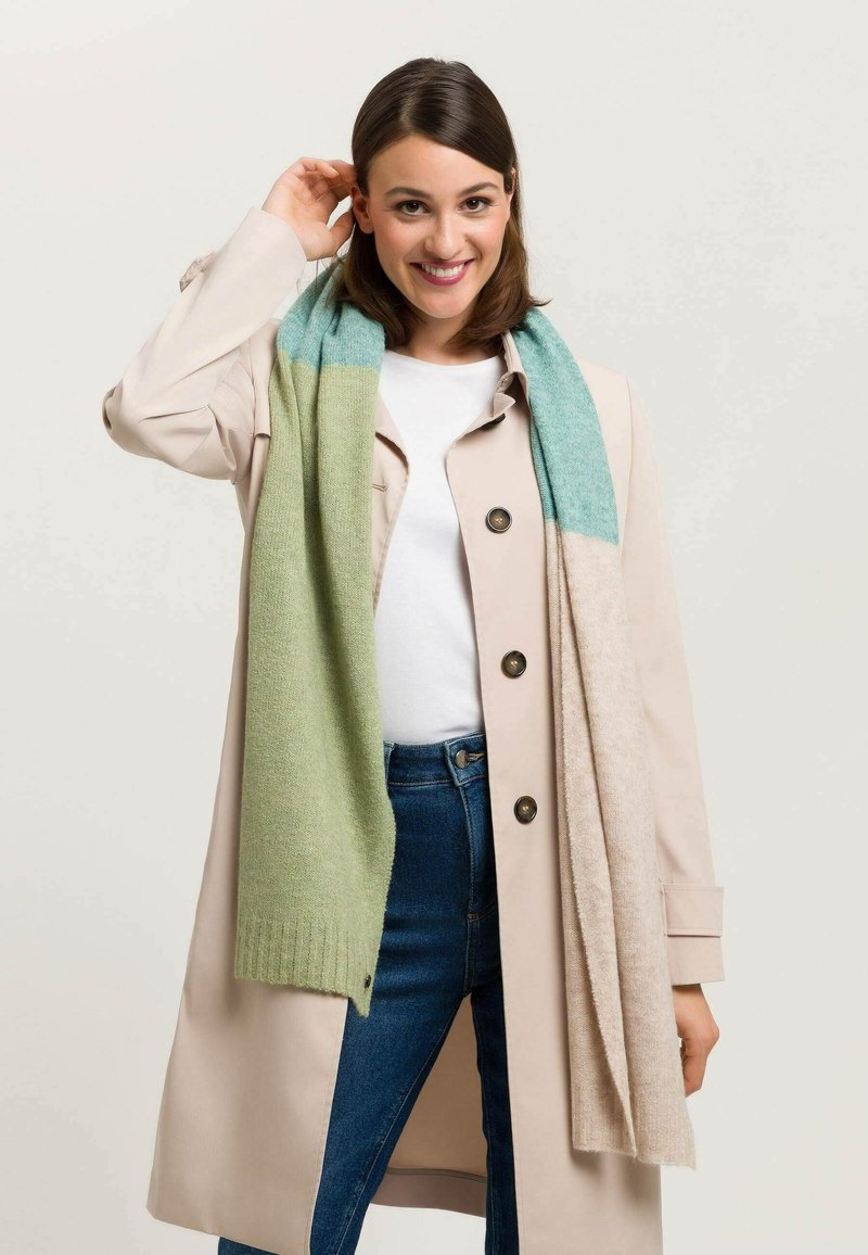 Écharpe en tricot avec un design dégradé en vert clair et turquoise, au toucher doux, drapée de manière décontractée sur un manteau crème.