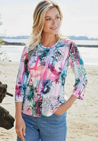 Bunt gemusterte Bluse mit floralem Design, Rundhalsausschnitt und Dreiviertelärmeln, kombiniert mit hellblauen Jeans. Lässige Strandszene.