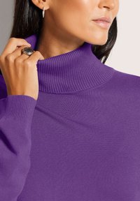 Frau trägt einen lila gerippten Rollkragenpullover, hält den Kragen mit der Hand und zeigt einen silbernen Ring, dazu trägt sie einen kleinen Hoops Ohrstecker.