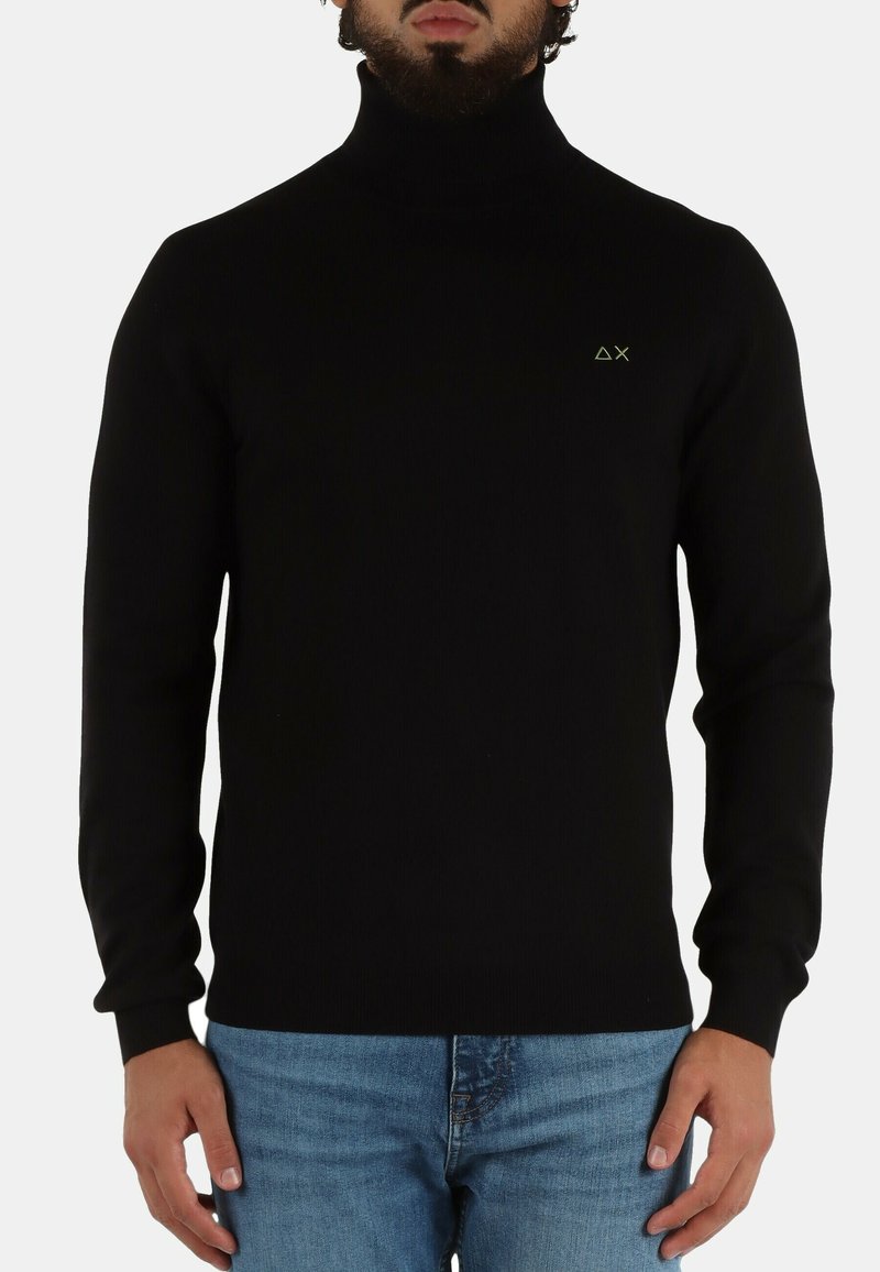 Maglione nero a collo alto con texture a costine, dotato di un piccolo logo verde sul petto. Abbinato a jeans azzurri chiari.