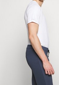 Homme portant une chemise blanche ajustée à manches courtes et un pantalon navy bleu avec cordon de serrage, se tenant de profil contre un fond uni.