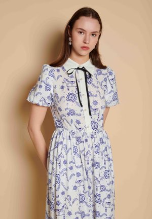 Jonge vrouw die een witte jurk draagt met blauwe bloemen- en paardenpatronen, pofmouwen, knoopsluiting aan de voorkant, een zwart lintje en pareloorbellen.