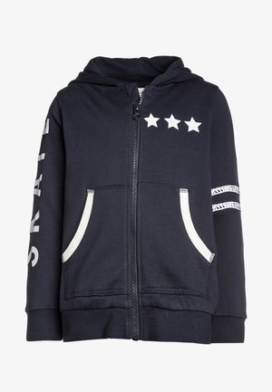 Haut à zip bleu marine avec trois étoiles blanches imprimées au-dessus de la fermeture éclair, présentant des rayures blanches sur les manches et deux poches avant.