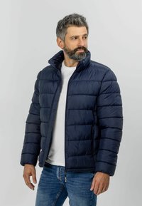 Veste en duvet navy avec un col montant, un design matelassé et des poches latérales zippées. Portée par-dessus un t-shirt blanc et associée à un jean bleu.