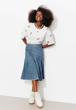 Femme souriante aux grands cheveux bouclés portant une chemise blanche à fleurs, une jupe midi en jean bleu, des chaussettes blanches et des baskets blanches, debout contre un mur blanc.